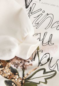 Kremasti duks s rebrastim manšetama, ukrašen cvjetnim uzorkom i metalik roza zlatnim šljokicama duž dizajna. Mekani materijal s teksturiranim završetkom.