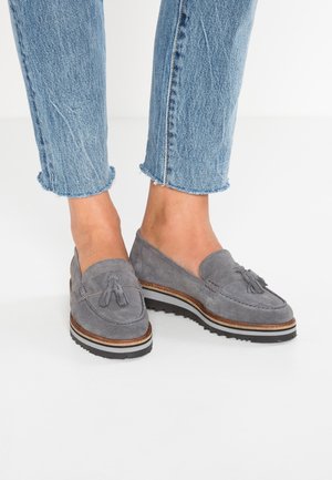 Šedé zamšové mokasíny s detailem třásní, vybavené nízkoprofilovou bílou pruhovanou podrážkou a klasickým mokasínovým designem. Nosit s roztrhanými džínami.