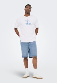 Ung mand iført en hvid T-shirt med en blå "Pensioneret" strandstolsgrafik, denimshorts, hvide sokker og hvide sko.
