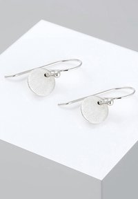 Pendientes circulares de plata con un acabado texturizado, que presentan un pequeño acento en forma de bola y ganchos alargados, exhibidos sobre un pedestal blanco.