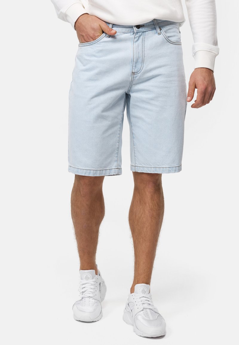Shorts en denim bleu clair à coupe décontractée et ourlet roulé, avec poches avant et fermeture éclair, portés avec des baskets blanches.