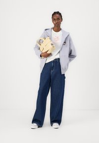 Sudadera con cremallera gris claro, camiseta gráfica blanca, jeans oscuros de denim sueltos y una pequeña bolsa amarilla. Sin patrones o herrajes notables.