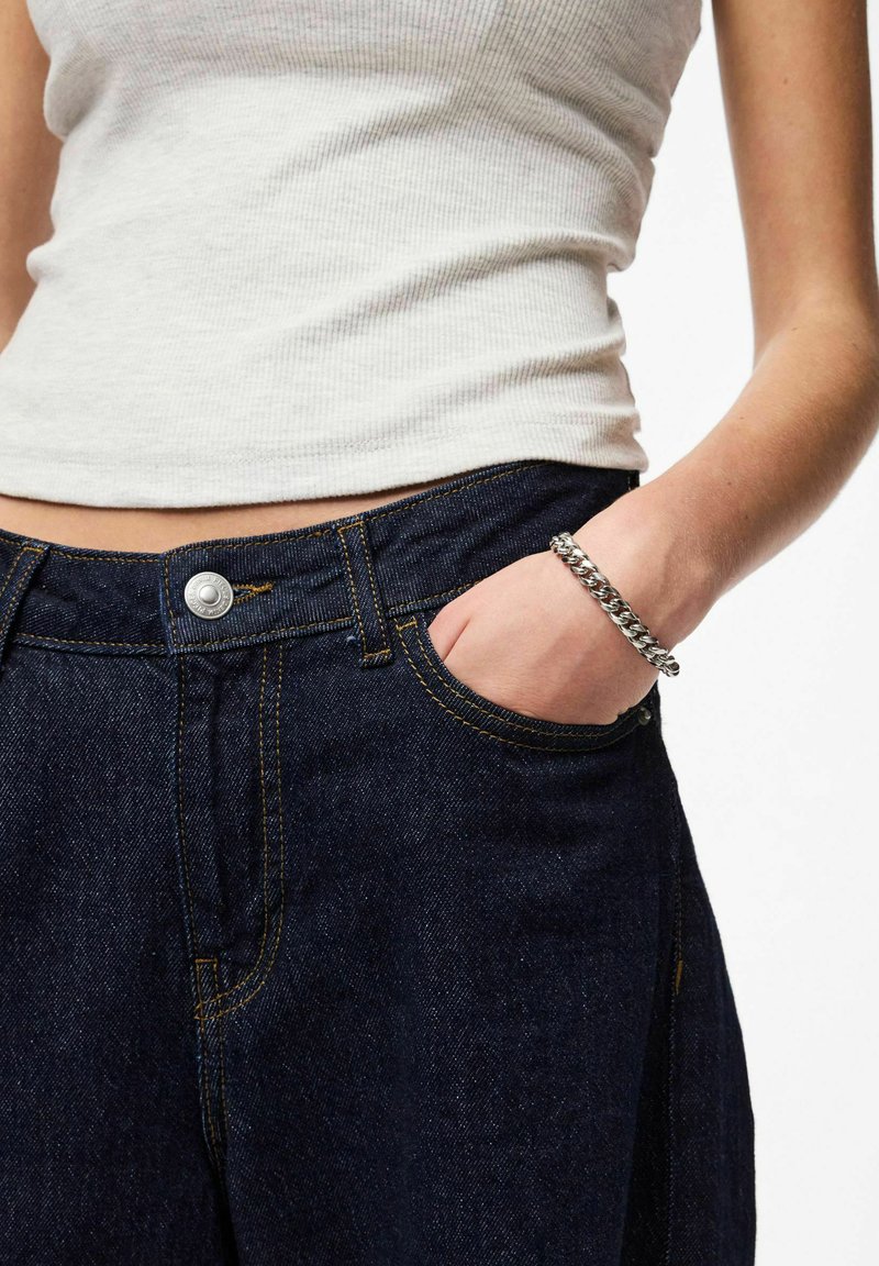 Jean en denim bleu foncé avec un motif de couture orange, doté d'une fermeture par bouton et d'une poche avant, associé à un débardeur côtelé gris clair et un bracelet argenté.