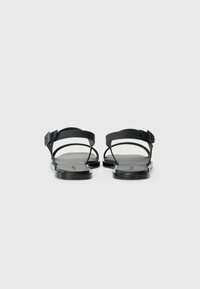 Black sandals with two thin straps and a buckle detail. Flat sole with a textured footbed. Minimalist design and sleek finish.