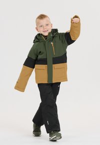 Kind in een groen, zwart en tangekleurde kleurafblok ski-jack met een capuchon en meerdere zakken, gecombineerd met zwarte broeken en groene schoenen.