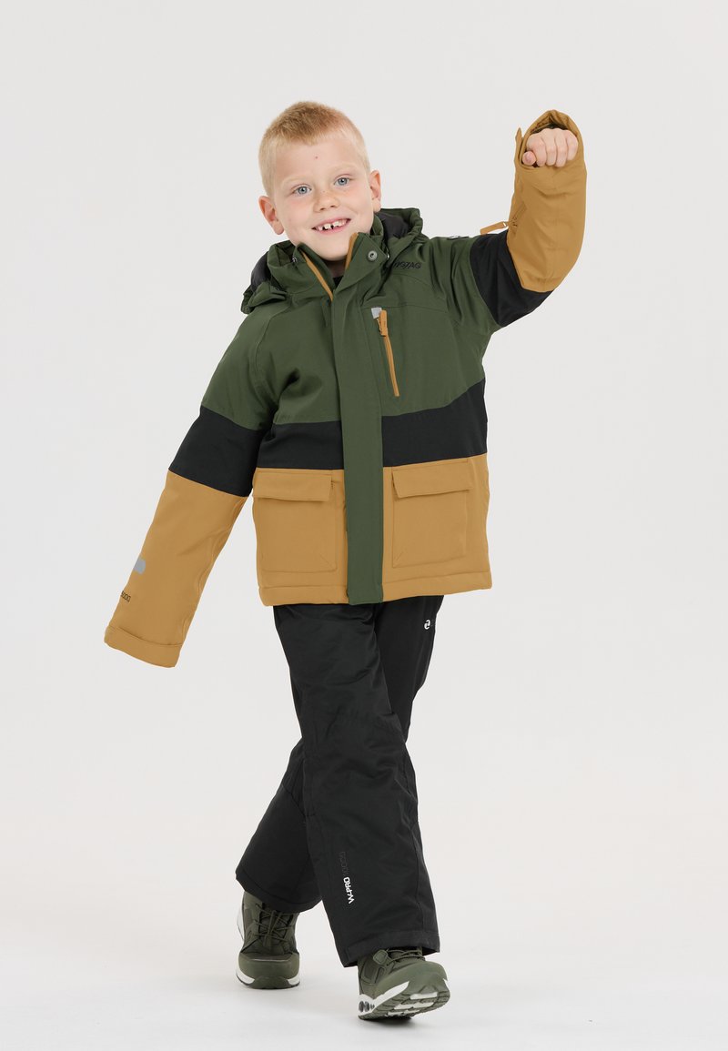Kind in een groen, zwart en tangekleurde kleurafblok ski-jack met een capuchon en meerdere zakken, gecombineerd met zwarte broeken en groene schoenen.