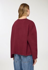 Felpa oversize color marrone rosso in cotone morbido, con scollo rotondo, spalle scese e orlo dritto. Indossata con jeans blu larghi.