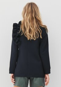 Crni blazer s detaljem u obliku nabora na ramenu, prilagođenog kroja, dužih rukava i glatke teksture tkanine. Noseni s zelenim šortsem.