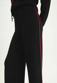 Pantalons de survêtement noirs avec une coupe ajustée, fabriqués en tissu doux, dotés d'une bande latérale rouge bien visible et d'une taille avec cordon de serrage réglable.