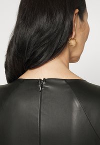 Robe en cuir noir avec un col haut, une texture lisse et une fermeture à glissière au dos. Comprend des boucles d'oreilles en anneau pour un détail supplémentaire.