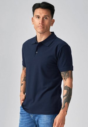 Muž s krátkymi tmavými vlasmi, oblečený v námornícky modrom polo tričku a svetlomodrých džínsoch, ukazujúci tetovania na oboch predlaktiach, stojaci na sivom pozadí.