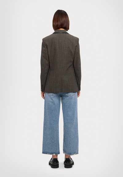 Femme de dos, portant un blazer gris foncé, un jean large bleu clair et des chaussures à plateforme noires, sur fond blanc.