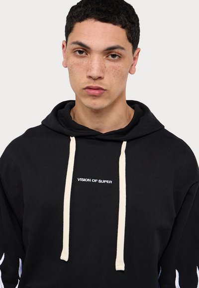 Sudadera negra con bolsillo frontal, cordones en color crema y logotipo "VISION OF SUPER" en blanco. Presenta patrones de llamas en las mangas.