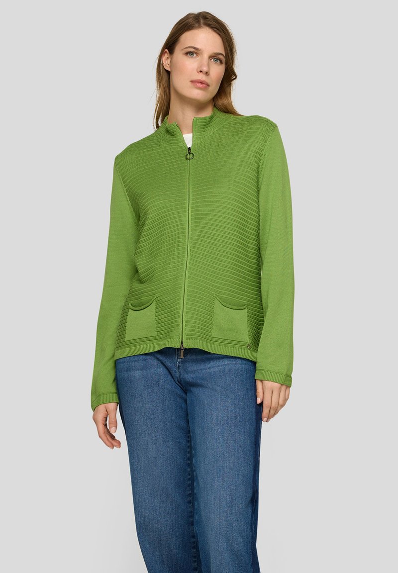 Grüne Strickjacke mit Reißverschluss, geripptem Muster, Stehkragen und zwei Vorderentaschen, kombiniert mit blauen Jeanshosen.