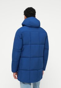 Modrá prošívaná bunda typu puffer s velkou kapucí, která má vertikální švy a nášivku s logem na levém rukávu. Texturovaný, zateplený design.