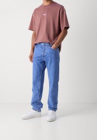 Camiseta de algodón marrón con un pequeño logo, combinada con unos jeans de mezclilla azules y zapatillas blancas. Los jeans tienen un corte relajado con costuras visibles.