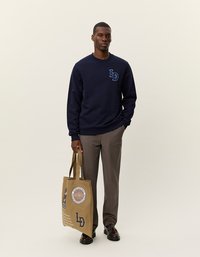 Sötétkék pulóver hímzett "LD" logóval, bézs nadrággal párosítva. A modellen egy barna vászontáska van, grafikai nyomtatással.