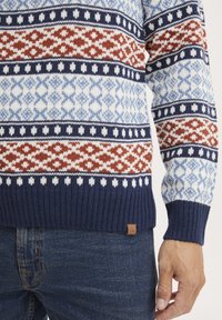 Kötött pulóver kék és fehér geometrikus mintával, piros részletekkel. bordázott mandzsettákkal és aljjal. Barna logó matrica alul.