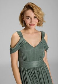 Robe de soirée plissée verte avec des bretelles décolletées, encolure en V et taille ajustée. Texture scintillante avec des lignes verticales.