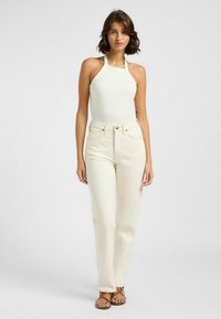 Top corto blanco de canalé con cuello halter, combinado con jeans de tiro alto en color crema. Presenta un diseño ajustado y una textura suave. Sandalias marrones con tiras completan el outfit.