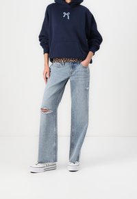 Marinblå hoodie med bågebroderi, tillsammans med ljusblå ripped jeans, prydda med ett leopardmönstrat bälte och vita canvas sneakers.