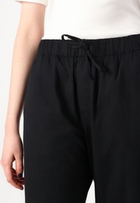 Pantalons noirs avec taille élastique et cordon de serrage, présentant une texture lisse et une coupe droite, montrés juste au-dessus du niveau de la taille.