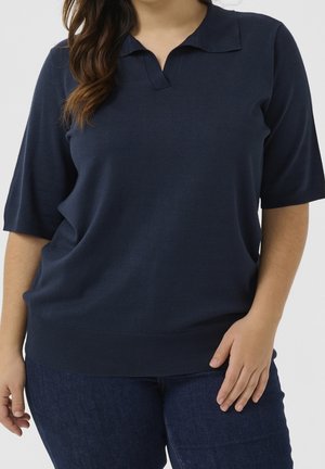 Kobieta ubrana w granatową krótko rękawową dzianinową koszulkę polo i ciemnoniebieskie jeansy, stojąca z jedną ręką spoczywającą na udzie.