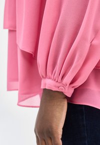 Σκούρο δέρμα χεριού που φοράει σκοτεινό παντελόνι, με το μανίκι ενός ροζ πουκάμισου που διαθέτει μανσέτα με κουμπί και ρ υτιδωμένο ύφασμα.