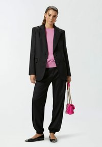 Giacca nera abbinata a un maglione rosa, pantaloni neri larghi e ballerine nere. Si tiene in mano una piccola borsa rosa con tracolla a catena.