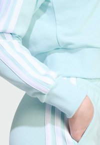 Sudadera verde menta clara con puños acanalados, que presenta rayas blancas y menta más clara a lo largo de las mangas, y un detalle de bolsillo en la parte delantera.