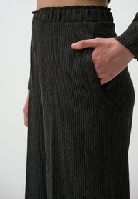 Pantalons noirs à jambes larges en tissu côtelé texturé, dotés d'une taille élastique et d'une poche sur le côté, présentés de la taille à mi-cuisse.