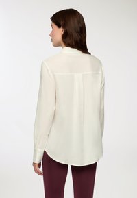 Blusa lunga con maniche a sbuffo di colore crema, con taglio dritto, polsini con bottoni, colletto moderato e tessuto liscio e leggero. La parte posteriore presenta una cucitura orizzontale.