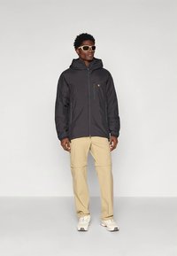 Crna jakna na patent s kapuljačom, sa logotipom i dva bočna džepa. Bež hlače sa zatvaračima, uparene sa svijetlim tenisicama i sunčanim naočalama.