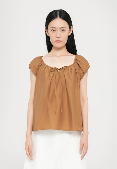 Femme aux longs cheveux noirs portant un blouse courte et ample marron avec un nœud à l'avant et un pantalon blanc, se tenant contre un fond uni.