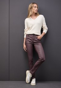 Crème, geribbelde trui met V-hals; bordeaux, slim-fit broek; lichtkleurige, sportieve sneakers; model leunend tegen een grijze muur.
