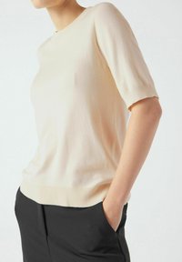 Maglione in maglia beige chiaro con maniche corte, scollo rotondo e orlo a coste. Indossato sopra pantaloni neri, che mostrano una consistenza e una vestibilità lisce.