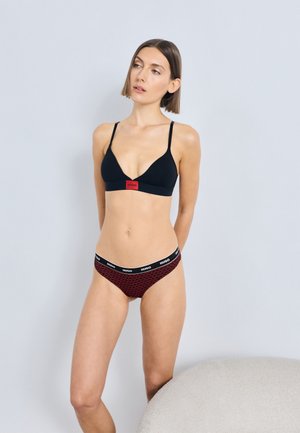 Μαύρο αδιάστατο σουτιέν με λεπτές τιράντες και κόκκινη ετικέτα με λογότυπο, συνδυασμένο με κόκκινα εσώρουχα με σχέδιο και μαύρη ζώνη μέσης με λογότυπο.