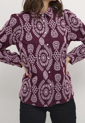 Frau trägt ein lila Langarmshirt mit hellrosa filigraner Blumenstickerei und schwarzen Hosen, steht mit den Händen in die Hüften gestützt.