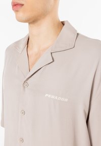Kortærmet, lys beige skjorte med knapper, blød tekstur, notch krave, og "PEGADOR" broderet i hvidt på brystet.