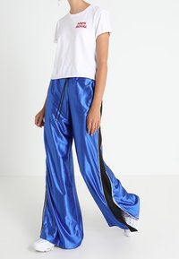 Persona con una camiseta blanca "100% ROYAL", pantalones anchos brillantes de color azul con rayas negras y zapatillas blancas, caminando contra un fondo simple.