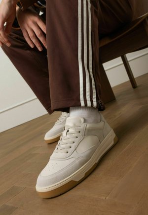 Witte leren sneakers met suède accenten, platte veters en een lichtbruine rubberen zool. Gecombineerd met bruine trainingsbroek met witte strepen.