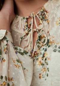 Blouse fleurie en tissu léger et transparent avec un motif à pois, dotée d'un décolleté ouvert noué, de manches amples et d'accents floraux pastel.