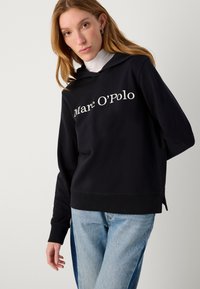 Schwarzer Kapuzenpullover mit weißem Text "Marc O'Polo", seitlichen Schlitzen und gerippten Bündchen. Kombiniert mit hellblauen Jeans. Glatte Textur.
