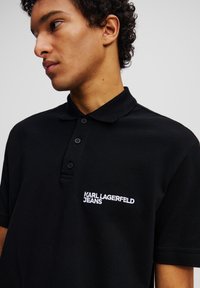 Μαύρο πόλο μπλουζάκι από υφασμάτινη υφή, με κλασικό γιακά και τρία κουμπιά. Σε εμφανές σημείο βρίσκεται το λευκό κεντημένο λογότυπο στο αριστερό στήθος.