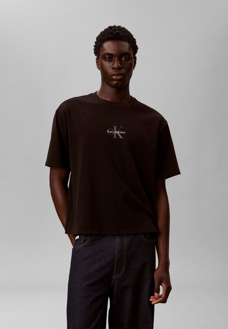 Νέος άνδρας που φοράει μαύρο T-shirt Calvin Klein και σκούρο τζιν, στέκεται μπροστά σε ένα απλό ανοιχτό γκρι φόντο.