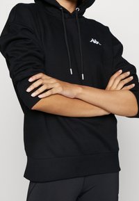 Persona con un hoodie nero e un logo bianco sul petto, braccia incrociate, maniche leggermente rialzate, in piedi contro uno sfondo semplice.