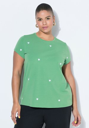 Kobieta z krótkimi włosami, ubrana w zielony t-shirt z małymi białymi serduszkami i czarne spodnie, stoi na tle białego tła.