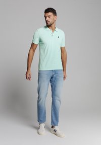Licht mintgroene polo met een drieknopsluiting, gedragen met lichtblauwe jeans en witte sneakers met marineblauwe accenten.