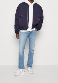 Navy bomber jacket with zippered pockets, worn over a white hoodie. Light blue jeans and white sneakers complete the outfit.