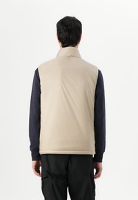 Beige Steppweste mit hohem Kragen, die eine glatte Textur, einen Reißverschluss vorne und schwarze Ärmel aufweist. Die Rückansicht zeigt minimale Designdetails.
